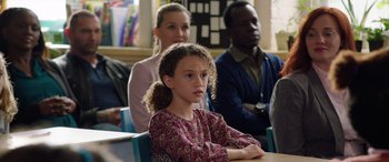 Movie still from “My Spy” (2020), directed by Peter Segal – A young girl sitting in front of a group of people in a classroom; Close Up shot, Over the shoulder angle