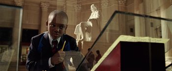 Movie still from “National Treasure” (2004), directed by Jon Turteltaub – A man in a suit and tie writing on a piece of paper in front of a statue; Medium shot, High angle