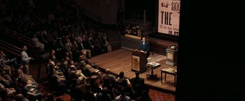 Movie still from “National Treasure: Book of Secrets” (2007), directed by Jon Turteltaub – A man standing at a podium in front of an audience; Extreme Wide shot, High angle