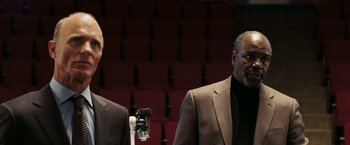 Movie still from “National Treasure: Book of Secrets” (2007), directed by Jon Turteltaub – Two men in suits are standing next to each other in front of an audience; Medium shot, Low angle