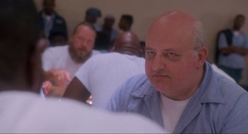 Movie still from “Natural Born Killers” (1994), directed by Oliver Stone – A man with a bald head is sitting next to a group of men; Close Up shot, Over the shoulder angle
