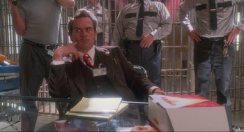 Movie still from “Natural Born Killers” (1994), directed by Oliver Stone – A man sitting at a table in front of a jail cell; Medium shot, Over the shoulder angle