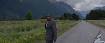 Movie still from “Needle in a Timestack” (2021), directed by John Ridley – A man in a gray suit standing next to the side of a road; Medium shot, Low angle