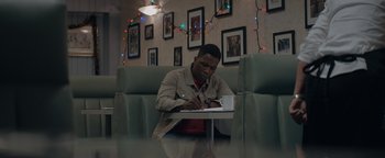 Movie still from “Needle in a Timestack” (2021), directed by John Ridley – A man sitting at a table writing on a notepad; Medium shot, Low angle
