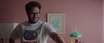 Movie still from “Neighbors” (2014), directed by Nicholas Stoller – A man in a white t - shirt is standing in front of a pink wall; Medium shot, Over the shoulder angle