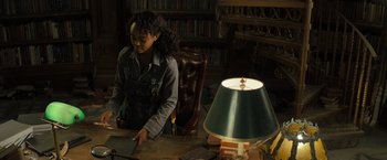 Movie still from “Nightbooks” (2021), directed by David Yarovesky – A woman sitting at a desk in front of a lamp; Medium shot, High angle