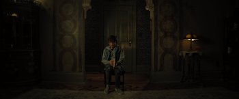 Movie still from “Nightbooks” (2021), directed by David Yarovesky – A person sitting on a chair holding a book in a room; Wide shot, High angle