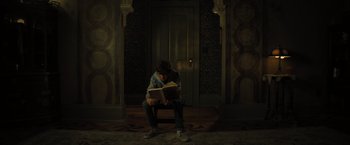 Movie still from “Nightbooks” (2021), directed by David Yarovesky – A man sitting on a chair reading a book; Wide shot, High angle