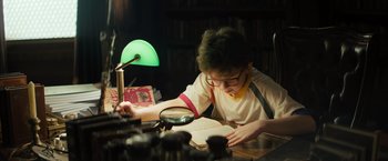 Movie still from “Nightbooks” (2021), directed by David Yarovesky – A boy is reading a book with a magnifying glass; Medium shot, High angle