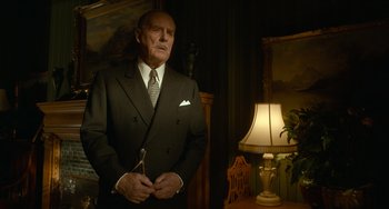 Movie still from “Nightmare Alley” (2021), directed by Guillermo del Toro – An older man wearing a suit and tie standing in front of a lamp; Medium shot, Low angle