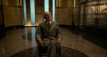 Movie still from “Nightmare Alley” (2021), directed by Guillermo del Toro – An older man in a suit and tie sitting in a chair in front of a clock tower; Medium shot, Low angle
