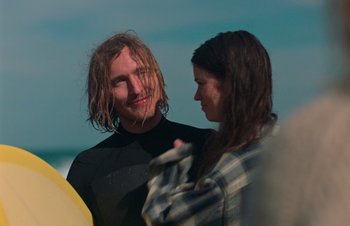 Movie still from “Nitram” (2021), directed by Justin Kurzel – A man and a woman standing next to a surfboard on the beach; Close Up shot, Over the shoulder angle
