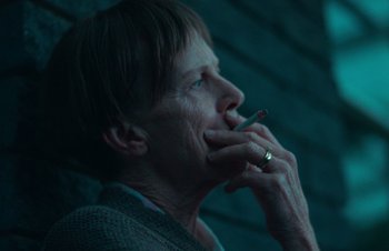 Movie still from “Nitram” (2021), directed by Justin Kurzel – An older woman smoking a cigarette in a dark room; Close Up shot, High angle