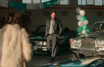 Movie still from “Nitram” (2021), directed by Justin Kurzel – A man in a suit and tie walking in front of some cars; Wide shot, Over the shoulder angle