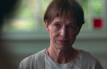 Movie still from “Nitram” (2021), directed by Justin Kurzel – An older woman with short brown hair wearing a white shirt; Close Up shot, Over the shoulder angle