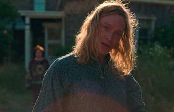 Movie still from “Nitram” (2021), directed by Justin Kurzel – A man with long blonde hair standing in front of a building; Medium shot, Over the shoulder angle