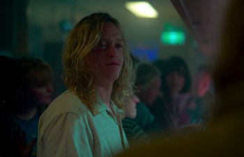 Movie still from “Nitram” (2021), directed by Justin Kurzel – A man with blonde hair is standing in front of a group of people; Medium shot, Low angle