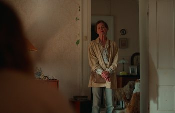 Movie still from “Nitram” (2021), directed by Justin Kurzel – An older woman standing in a living room holding a wii controller; Medium shot, Over the shoulder angle