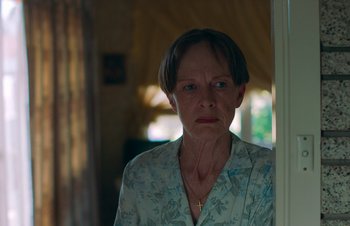 Movie still from “Nitram” (2021), directed by Justin Kurzel – An older woman standing in front of a window; Close Up shot, Over the shoulder angle