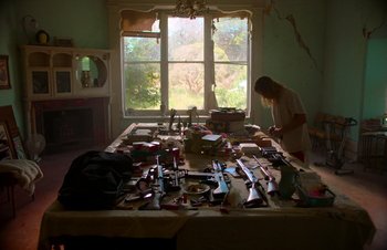 Movie still from “Nitram” (2021), directed by Justin Kurzel – A woman standing in front of a table covered with many objects; Wide shot, High angle