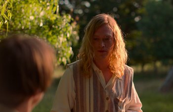 Movie still from “Nitram” (2021), directed by Justin Kurzel – A man with long blonde hair standing in a field; Medium shot, Over the shoulder angle