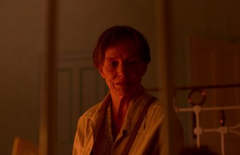 Movie still from “Nitram” (2021), directed by Justin Kurzel – An older woman is standing in a dimly - lit room; Close Up shot, Over the shoulder angle