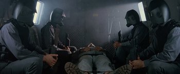 Movie still from “No Escape” (1994), directed by Martin Campbell – A group of people sitting on the ground in a dark room; Medium shot, Low angle