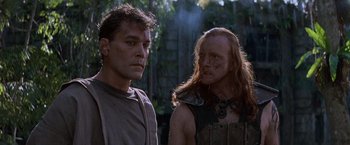 Movie still from “No Escape” (1994), directed by Martin Campbell – A man standing next to another man in a forest; Close Up shot, Low angle