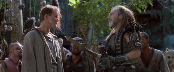 Movie still from “No Escape” (1994), directed by Martin Campbell – Two men in costume talking to each other in a forest; Medium shot, Over the shoulder angle