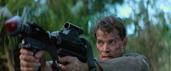 Movie still from “No Escape” (1994), directed by Martin Campbell – A person holding a gun in a forest; Close Up shot, Low angle