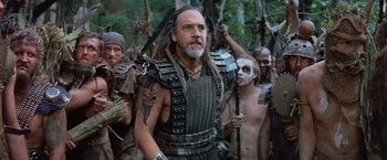 Movie still from “No Escape” (1994), directed by Martin Campbell – A group of people in costume in a forest; Medium shot, Low angle