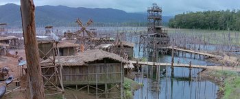 Movie still from “No Escape” (1994), directed by Martin Campbell – A group of wooden stilt houses in the middle of a body of water; Extreme Wide shot, High angle