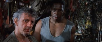 Movie still from “No Escape” (1994), directed by Martin Campbell – A man and a woman are standing next to each other; Close Up shot, Low angle