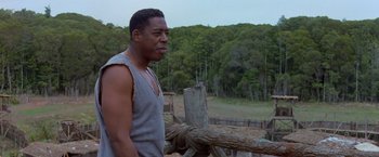 Movie still from “No Escape” (1994), directed by Martin Campbell – A man standing in front of a fence and trees; Medium shot, Low angle