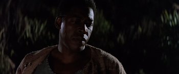 Movie still from “No Escape” (1994), directed by Martin Campbell – A man in a brown shirt is standing in the dark; Close Up shot, Low angle