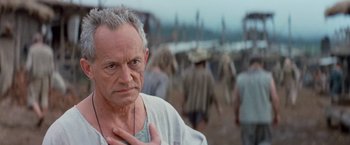 Movie still from “No Escape” (1994), directed by Martin Campbell – An older man in a white shirt is standing in front of a group of people; Close Up shot, Over the shoulder angle