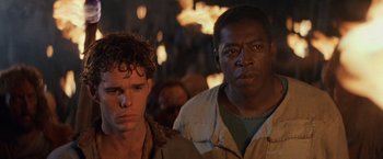 Movie still from “No Escape” (1994), directed by Martin Campbell – Two men standing next to each other in front of fire; Close Up shot, Over the shoulder angle