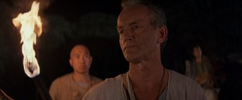 Movie still from “No Escape” (1994), directed by Martin Campbell – A man standing next to another man in a dark room; Close Up shot, Over the shoulder angle