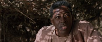 Movie still from “No Escape” (1994), directed by Martin Campbell – A person wearing a brown shirt near a tree; Close Up shot, Low angle