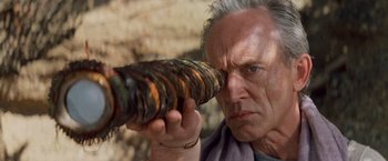 Movie still from “No Escape” (1994), directed by Martin Campbell – An older man looking through a tube of wood; Close Up shot, Low angle