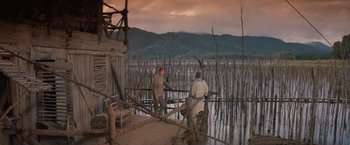 Movie still from “No Escape” (1994), directed by Martin Campbell – Two men standing in front of a body of water; Extreme Wide shot, High angle