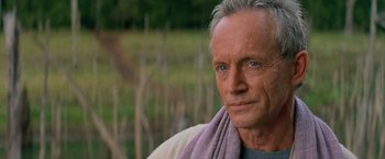 Movie still from “No Escape” (1994), directed by Martin Campbell – An older man with a towel around his neck; Close Up shot, Over the shoulder angle