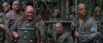 Movie still from “No Escape” (1994), directed by Martin Campbell – A group of men standing next to each other in a forest; Medium shot, Low angle