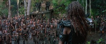 Movie still from “No Escape” (1994), directed by Martin Campbell – A group of men in a forest with a woman in the foreground; Wide shot, High angle