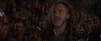 Movie still from “No Escape” (1994), directed by Martin Campbell – A man with dreads holding a cell phone in front of a group of people; Close Up shot, Low angle