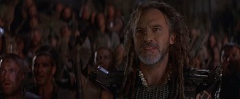 Movie still from “No Escape” (1994), directed by Martin Campbell – A man with dreads holding a book in a crowd of people; Close Up shot, Over the shoulder angle