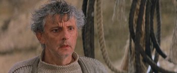Movie still from “No Escape” (1994), directed by Martin Campbell – An older man wearing a sweater and looking at the camera; Close Up shot, Low angle