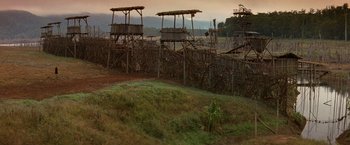 Movie still from “No Escape” (1994), directed by Martin Campbell – A grassy area next to a fence and a building; Extreme Wide shot, High angle