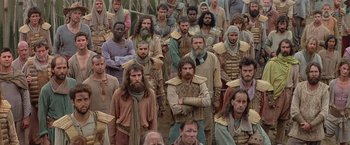 Movie still from “No Escape” (1994), directed by Martin Campbell – A large group of men standing together in a field; Wide shot, High angle