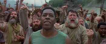Movie still from “No Escape” (1994), directed by Martin Campbell – A group of people in a field with a man in a green shirt; Close Up shot, Low angle